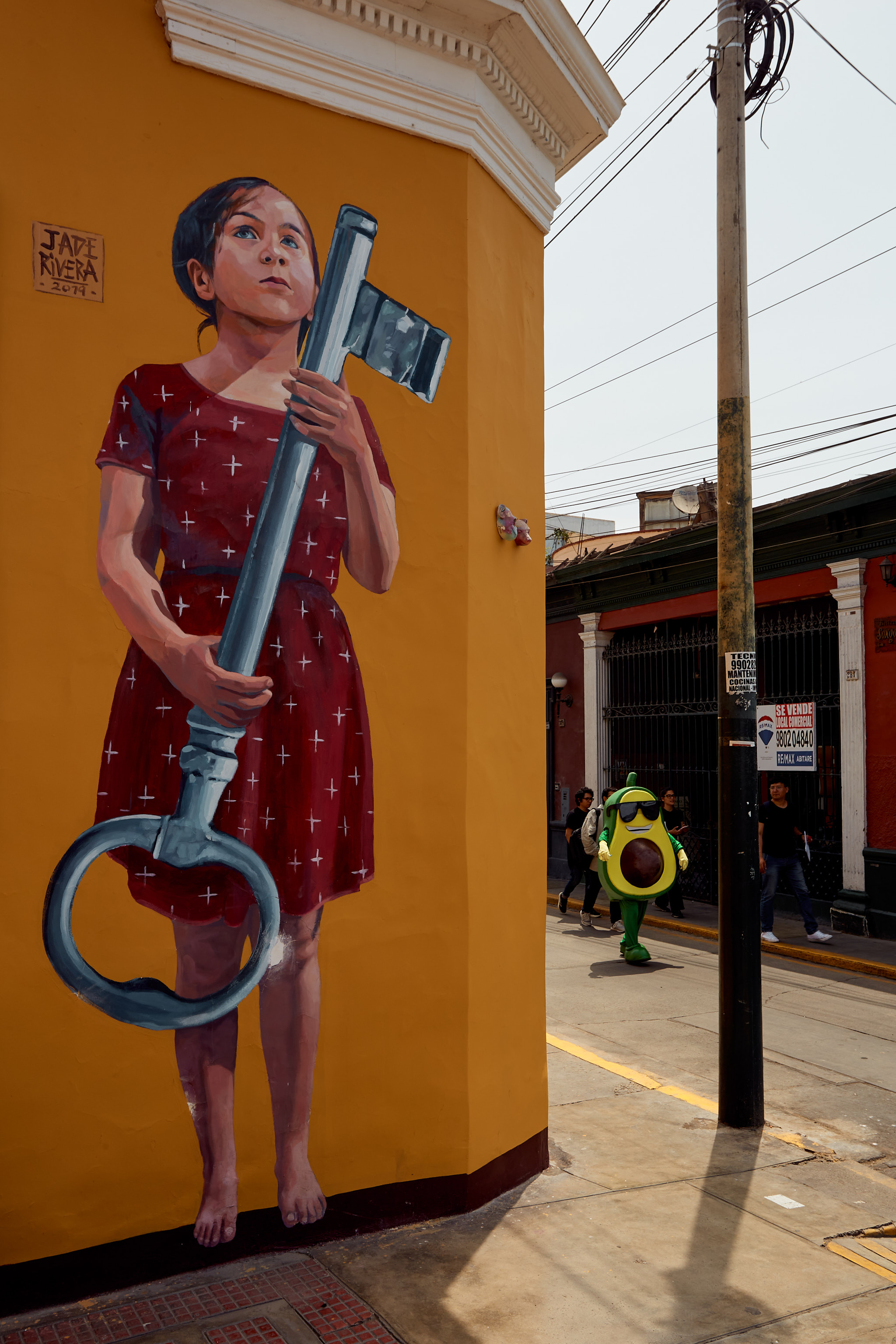 Mural s dívkou v červených šatech nesoucí velký klíč na žluté zdi v Barrancu, avokádový maskot v pozadí